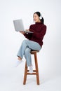 Young positive Asian woman in casual clothes wearing headphones and using her laptop on a chair Royalty Free Stock Photo