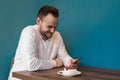 Young positive fellow European businessman in a white shirt spends time in a smartphone and smiles on a coffee break in a cafe Royalty Free Stock Photo
