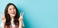 Young positive modern girl sending good vibes only, showing v-sign and smiling happy, standing against blue background Royalty Free Stock Photo