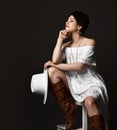 Young positive pretty short haired brunette woman in white elegant dress boots and hat sitting on stool looking aside Royalty Free Stock Photo