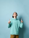 Young positive teen boy in blue hoodie and baseball cap standing pointing up with forefingers over pastel blue wall background