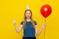 Young positive woman in a birthday cap holds red inflated helium balloon and shows thumb-up, feels very happy, stands over yellow