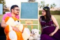 Young pregnant asian indian couple looking at each other sitting in a garden with lots teddy bear toys and blank chalkboard to Royalty Free Stock Photo