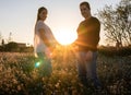 Young pregnant couple holding hands into white flowers field with the sunset and sun rays in the background Royalty Free Stock Photo