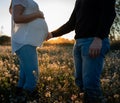 Young pregnant couple into white flowers field with the sunset and sun rays in the background