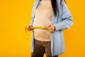 Young pregnant lady measuring her belly with tape to keep track of her fetus development, cropped, yellow background