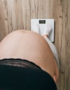 Young pregnant woman with big belly standing in white socks on digital scales on home living room laminate floor. health,