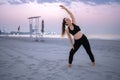 Young pregnant woman in black sportswear stretching on the sand beach with purple sunset over ocean in the background. Royalty Free Stock Photo