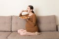 Young pregnant woman enjoys eating croissant on the sofa. Unhealthy pastry during pregnancy concept