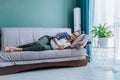 Young pregnant woman with glasses in white top, grey trousers and plaid shirt sleeping on grey sofa holding book at home