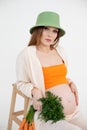 Young pregnant woman with long dark hair wear green bucket hat, sitting on chair, holding belly and bunch of carrots. Royalty Free Stock Photo