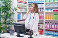 Young pregnant woman pharmacist talking on smartphone using computer at pharmacy