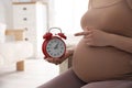 Young pregnant woman pointing at alarm clock near belly indoors, closeup. Time to give birth