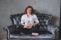 Young pregnant woman with red hair sitting on a gray sofa in the Baroque style. She`s wearing a white shirt, visible to