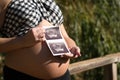 Young pregnant woman showing the ultrasound of her baby on her belly in an outdoor park. Concept pregnancy, maternity, baby, Royalty Free Stock Photo