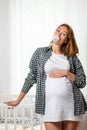 A young pregnant woman shows off her tummy and inflates a bubble of gum. White window in the background. Concept of pregnancy and