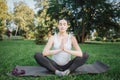 Young pregnant woman sit on yoga mate and pray outside in park. She keep hands close and sit in lotus pose. Model hold