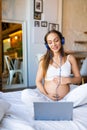 Young pregnant woman sits on a bed in blue headphones in front of a laptop monitor. She communicates - online video call Royalty Free Stock Photo