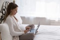 Young pregnant woman sits on bed with laptop