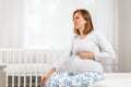 A young pregnant woman is sitting on the bed and writhing with pain in her belly. Children`s room in the background. The concept