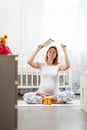 A young pregnant woman with a smile sits on the floor next to baby booties and holds a book over her head. Children`s room in the