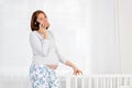 A young pregnant woman is standing in the nursery, leaning on a baby cradle and chatting on her smartphone. Concept of modern Royalty Free Stock Photo