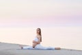 Young pregnant woman in white dress sitting on the beach near blue sea and breathing. Summer vacation during pregnancy Royalty Free Stock Photo