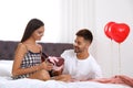 Young presenting gift to his girlfriend in bedroom decorated with air balloons. Celebration of Saint Valentine`s Day