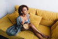 Young pretty african girl enjoying healthy breakfast, eating granola sitting on yellow couch, sofa, home morning Royalty Free Stock Photo