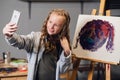 A young pretty artist takes a selfie against the background of her painting in the Studio.