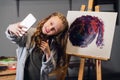 A young pretty artist takes a selfie against the background of her painting in the Studio.