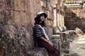 Young pretty asian girl dressed in a striped clothes and white hat sits on the foundation of an ancient hindu temple ruins and