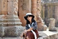 Young pretty asian girl dressed in a striped dress and white hat sits on the foundation of an ancient hindu temple. Prasat Hin