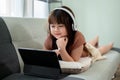 Young pretty Asian girl is studying online on her digital tablet while lying on a sofa Royalty Free Stock Photo