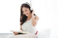 Young pretty Asian woman reading book and and listening music in modern headphones connected to smartphone on bed. Concepts of Royalty Free Stock Photo