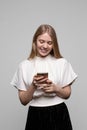 Young pretty blond girl with long hair smiling and using cell phone isolated on gray background. Modern communication Royalty Free Stock Photo