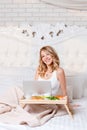 Young pretty blond woman sitting on bed with laptop having breakfast, freelancer or blogger at home. Woman works on Royalty Free Stock Photo