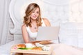 Young pretty blond woman sitting on bed with laptop having breakfast, freelancer or blogger at home. Woman works on Royalty Free Stock Photo