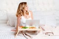 Young pretty blond woman sitting on bed with laptop having breakfast, freelancer or blogger at home. Woman works on Royalty Free Stock Photo