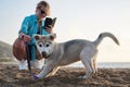 Young pretty blond woman, taking picture on her phone of her small husky puppy. Owner and the light grey dog, playing on the sandy