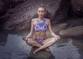 Young pretty blonde female wearing colorful bikini sitting on the rock and doing meditation yoga.