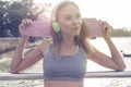 Young pretty blonde fitness female wearing bright green headphones listening music and holding pink skateboard while standing on