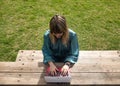 Young, pretty, blonde woman in green shirt, sitting on a wooden bench in a park, working with her laptop, view from above. Royalty Free Stock Photo