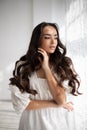 Young pretty brunette girl with incredible makeup in white summer dress standing near the window.