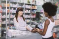 Young pretty Caucasian woman pharmacist helping a female African customer, dispensing her prescription medicine with a