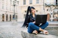 Young pretty concentrated Asian woman with brunette ponytail hairstyle, freelancer, sitting at old city fountain