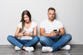 Young pretty couple typing on phone while sitting on the floor isolated on gray background
