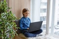 A young pretty emotional teenage girl is sitting on a cozy windowsill with pillows and a laptop. He looks at the monitor in Royalty Free Stock Photo