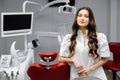 A young pretty female dentist is standing by the dental chair in the office and looking into the camera Royalty Free Stock Photo