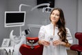 A young pretty female dentist is standing near the dental chair in the office, holding tools for work and looking into the camera Royalty Free Stock Photo